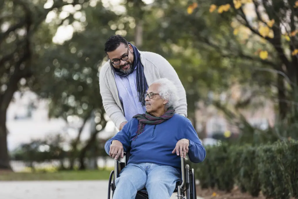 An Adult man pushing senior man in wheelchair while discussing what is respite care ? for seniors at Burcham Hills in East Lansing, MI.