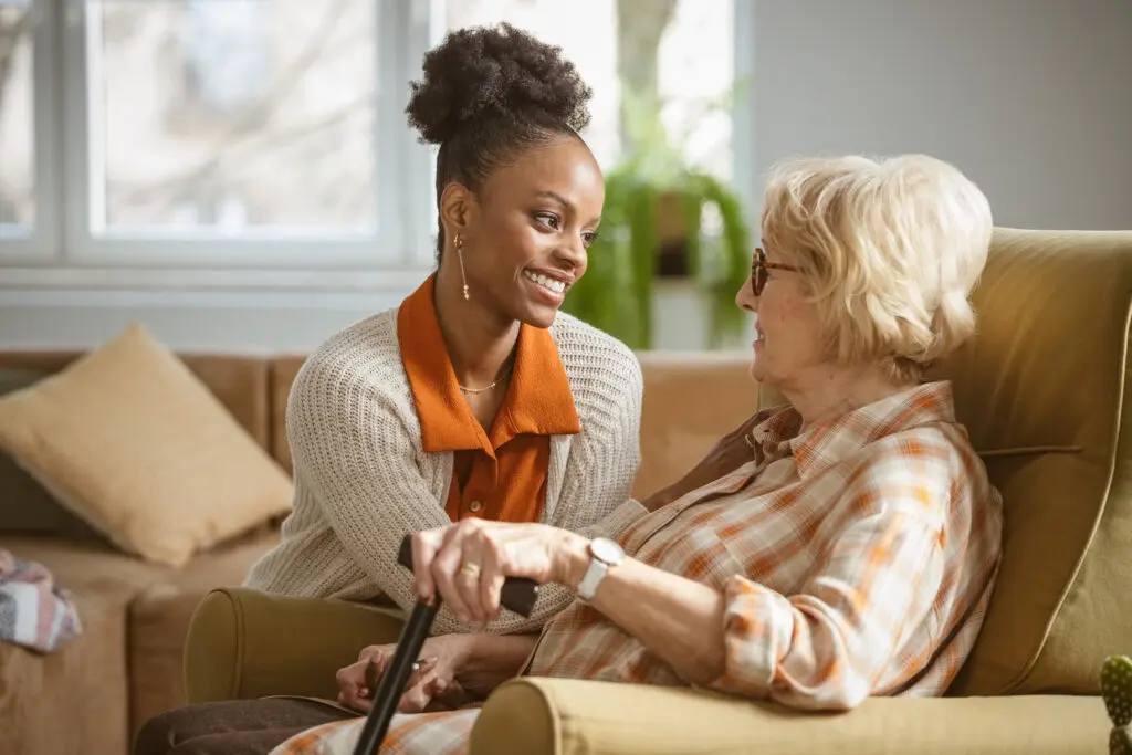 A woman caregiver kneeling next to senior woman in chair while talking about signs for Assisted Living at Burcham Hills in East Lansing, MI