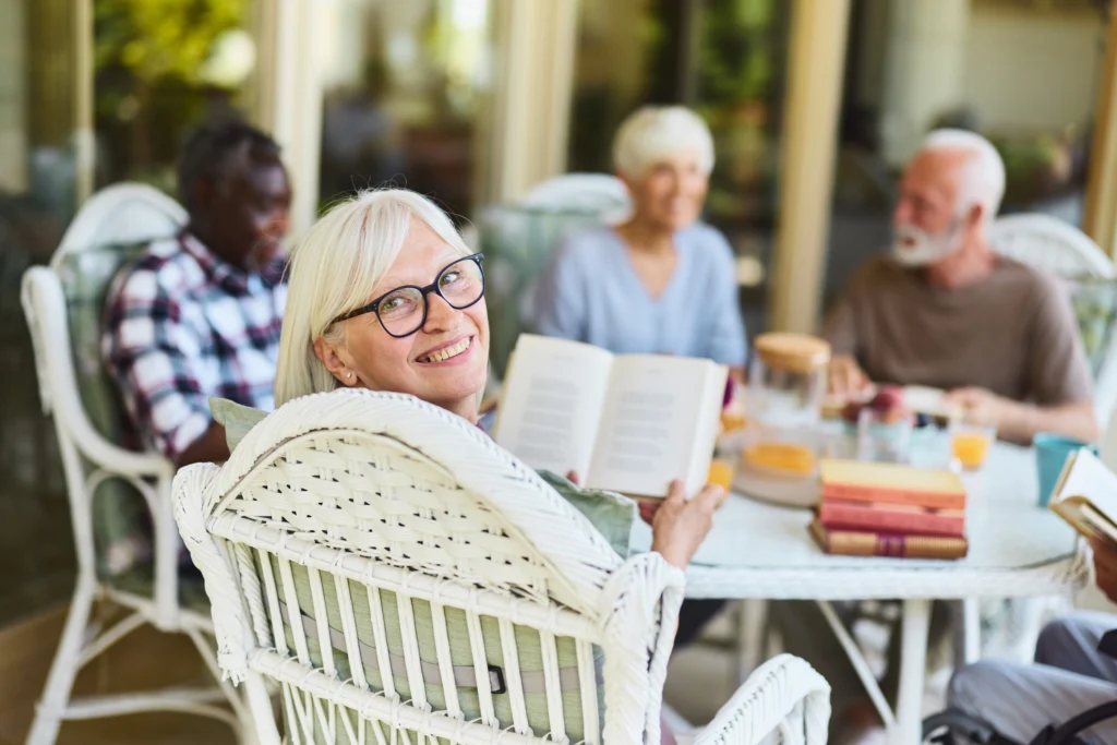 A senior woman smiling and reading a book on assisted living costs in Michigan. She is preparing to discuss assisted living and elderly costs with the 3 seniors at her table.