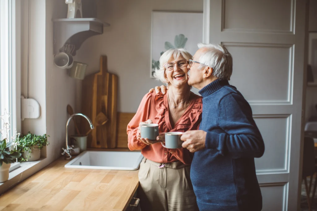 senior man kissing senior woman on the cheek in kitchen while holding coffee cups. They were discussing "What Is a CCRC?" from their home at Burcham Hill, a life plan community in East Lansing, MI.